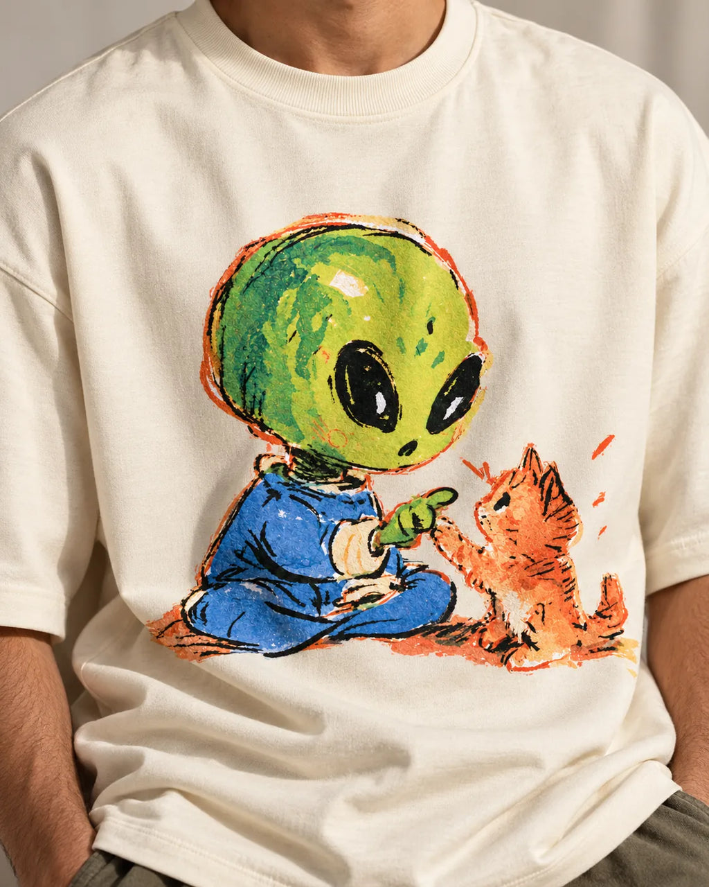 Close-up of a green alien touching paws with an orange kitten printed on an off-white oversized French terry t-shirt