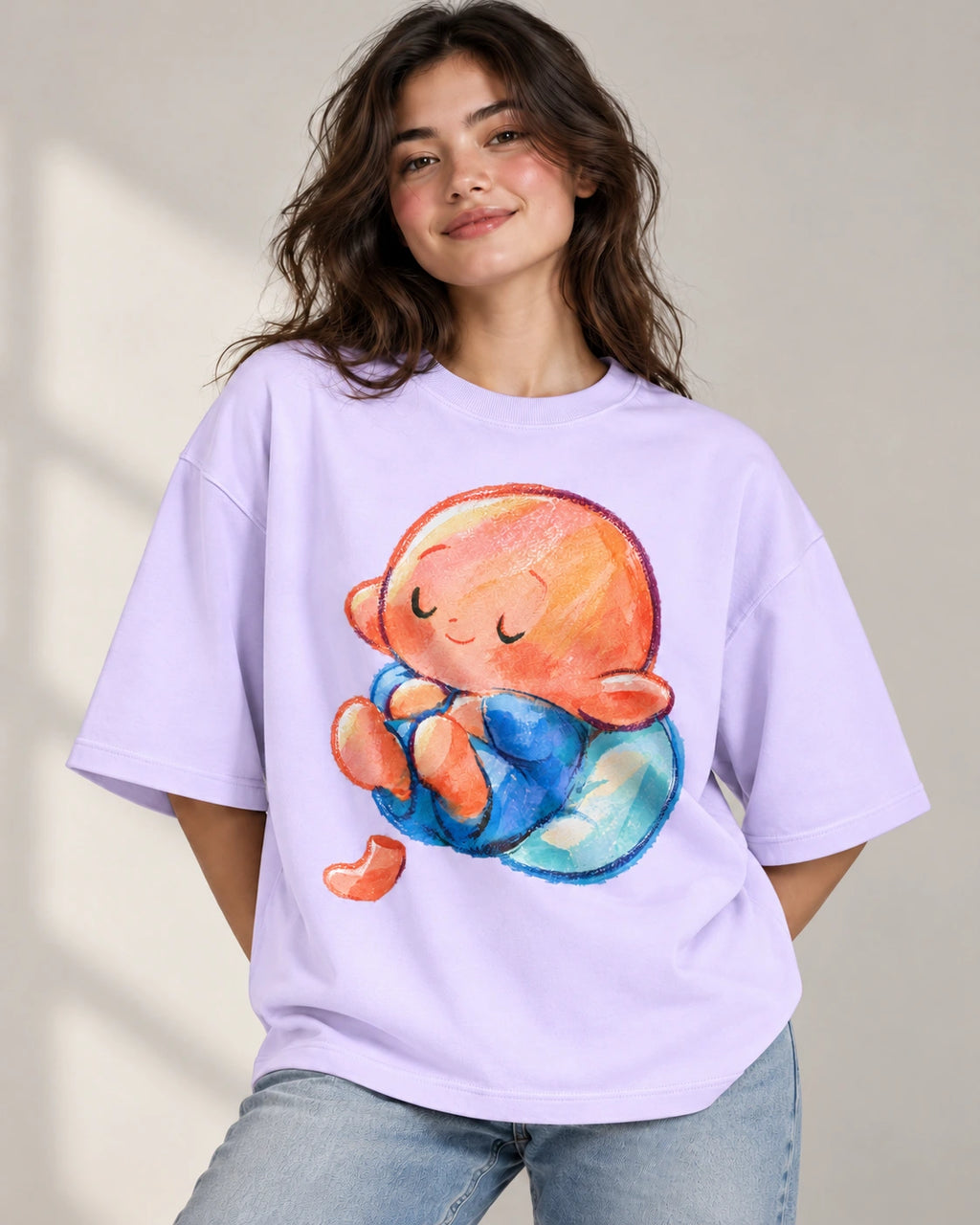 Woman in a lavender oversized French terry t-shirt printed with a sleeping orange-headed creature curled on a blue cushion