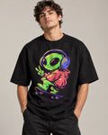 Man in a black oversized French terry t-shirt printed with a green alien in headphones skateboarding with a peace sign