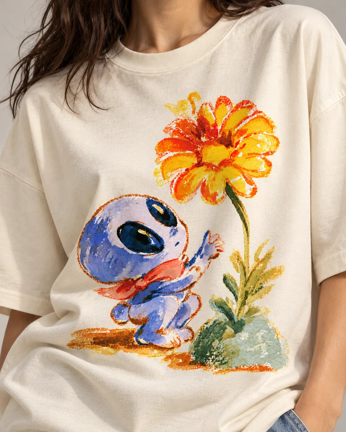 Close-up of a blue alien in a red scarf reaching for an orange flower printed on an off-white oversized French terry t-shirt