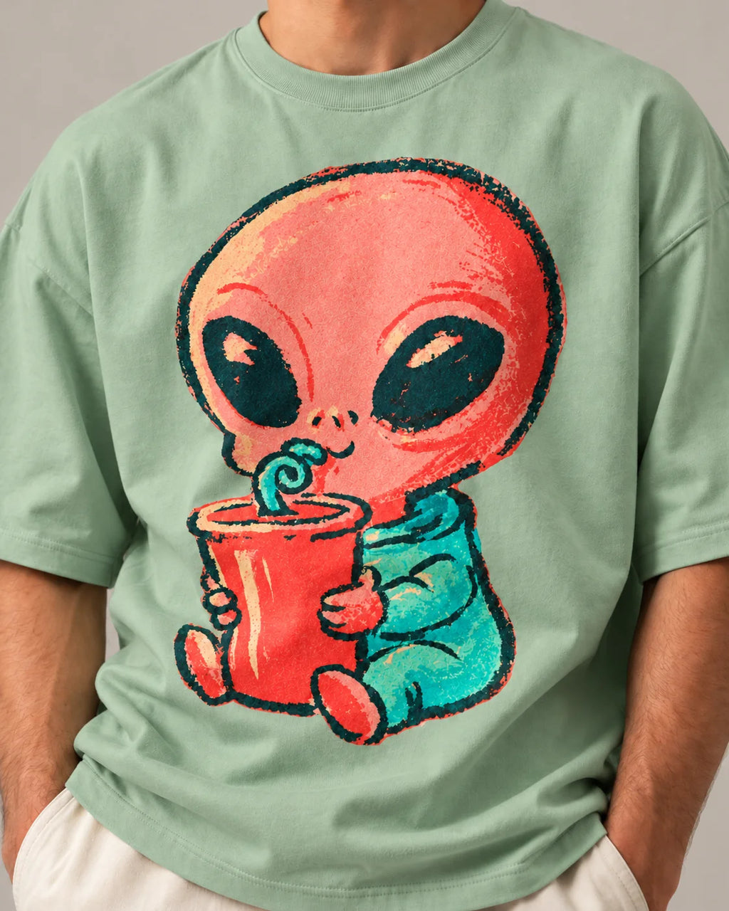 Close-up of a coral alien sipping a red cup through a curly straw printed on a jade oversized French terry t-shirt