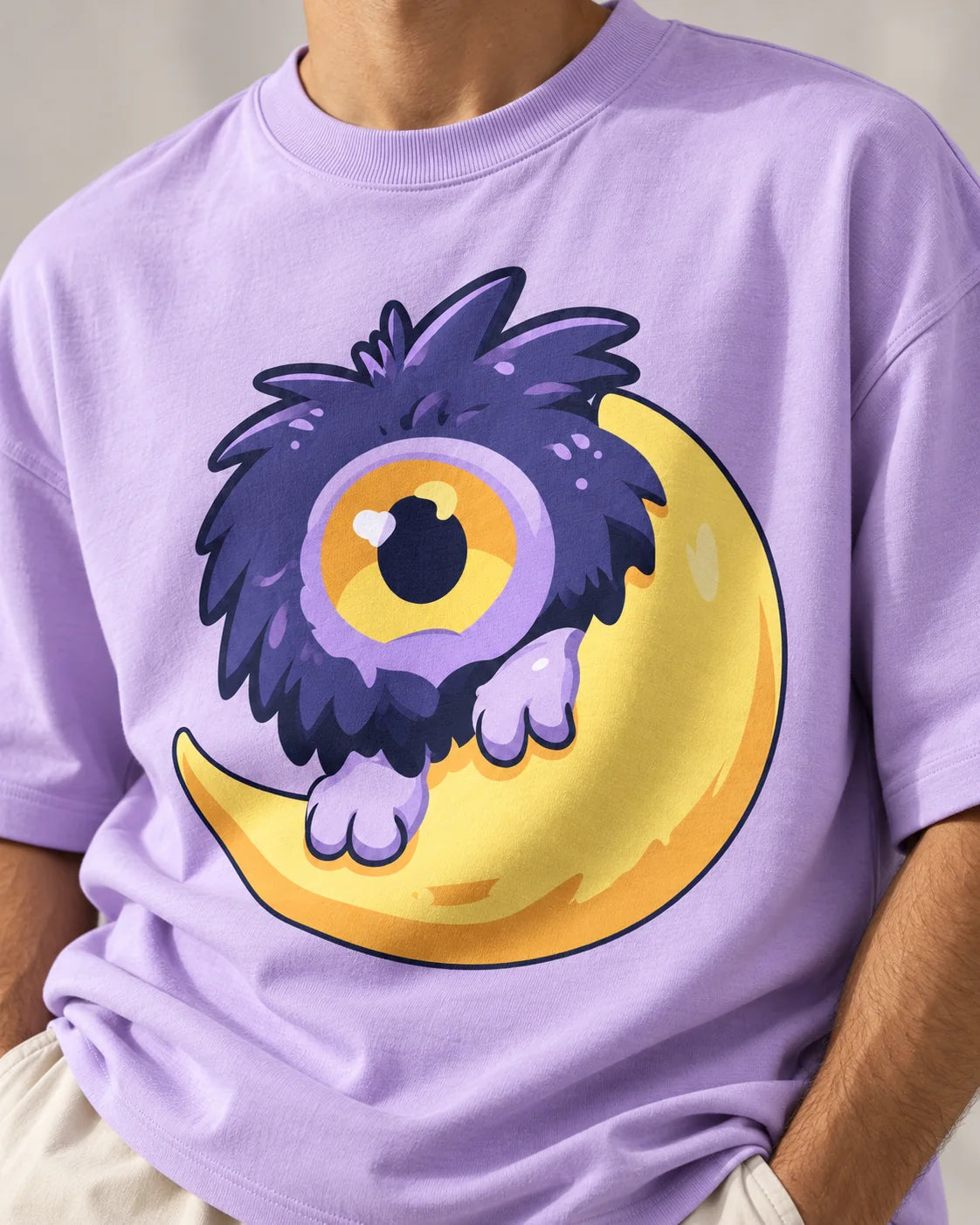 Close-up of a fluffy one-eyed purple monster on a yellow moon printed on a lavender oversized French terry t-shirt
