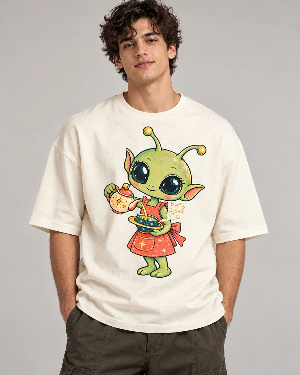 Man in an off-white oversized French terry t-shirt printed with a green alien in an apron pouring tea from a teapot