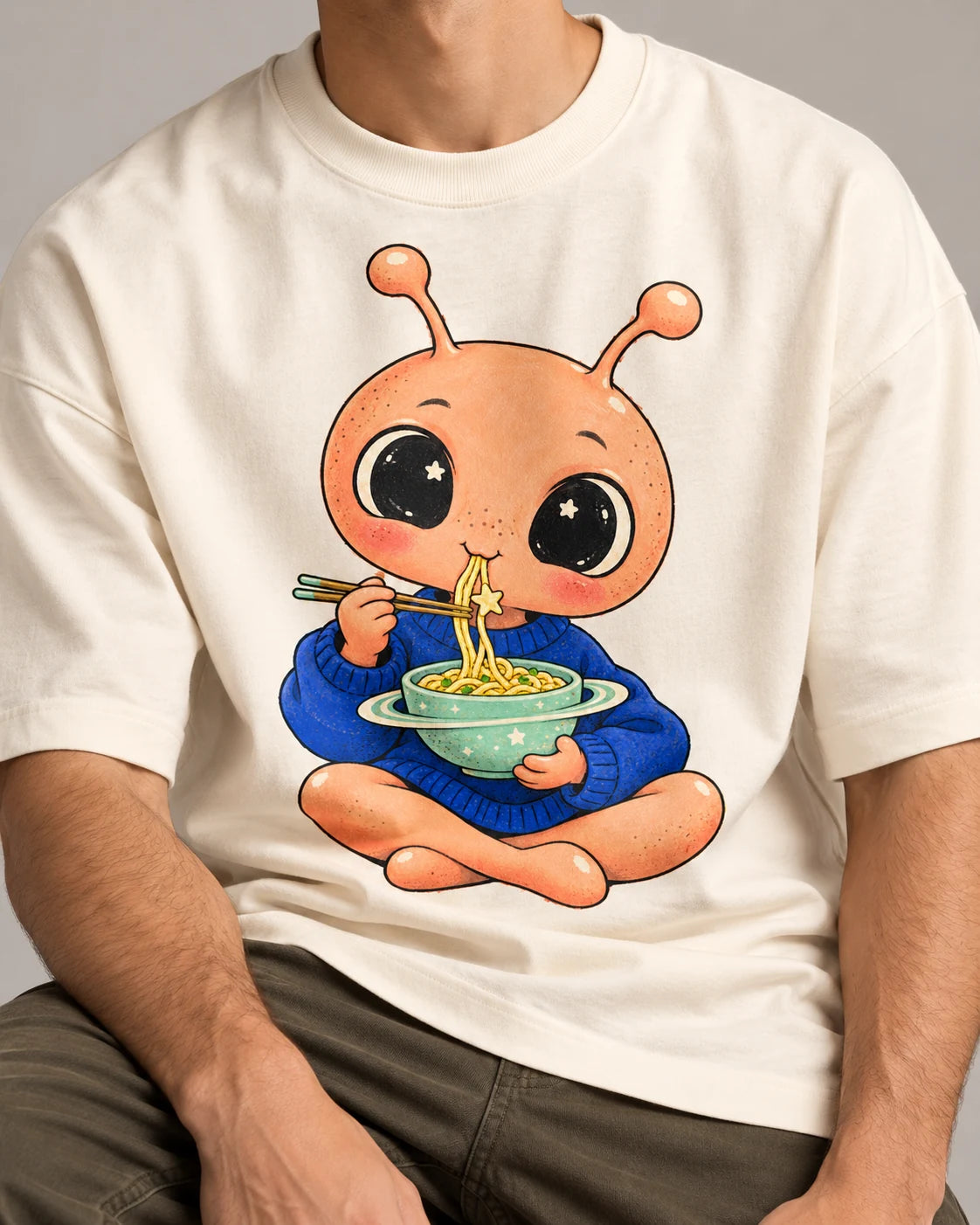 Close-up of a peach alien eating noodles from a ringed bowl printed on an off-white oversized French terry t-shirt