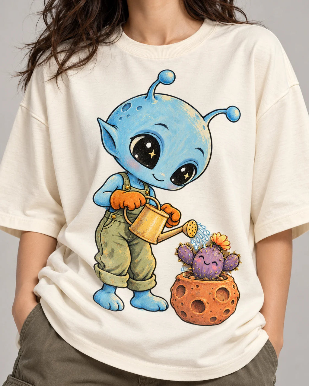 Close-up of a blue alien watering a cactus in a meteor-like pot printed on an off-white oversized French terry t-shirt