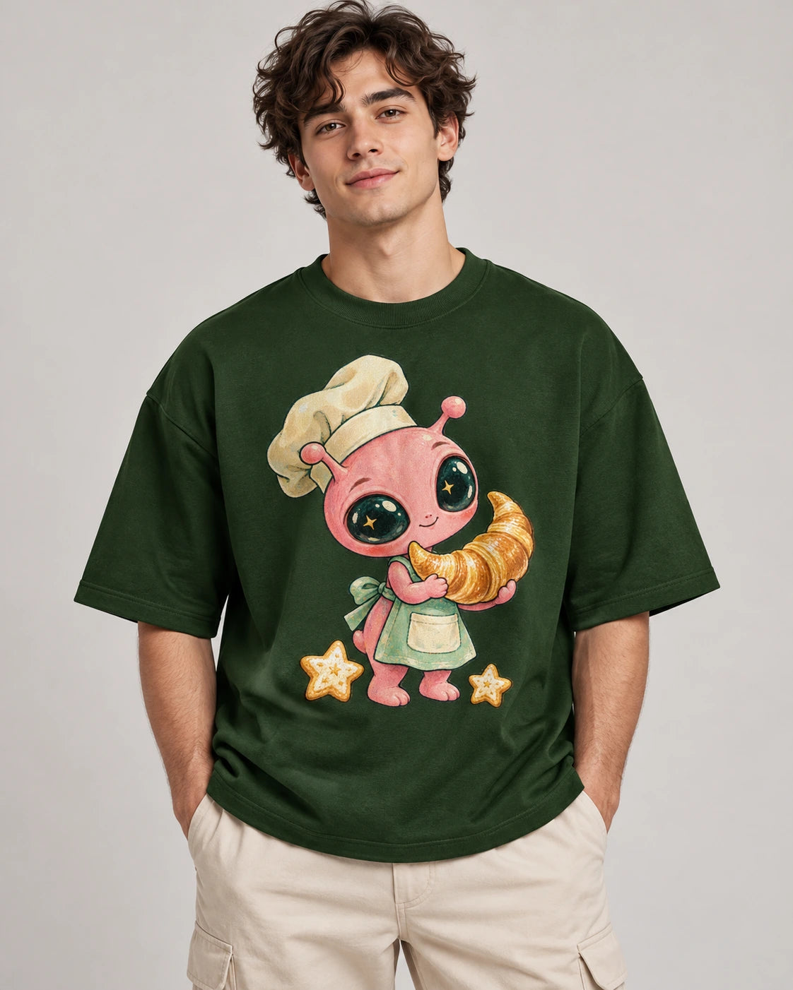 Man in a bottle green oversized French terry t-shirt printed with a pink alien chef carrying a giant croissant